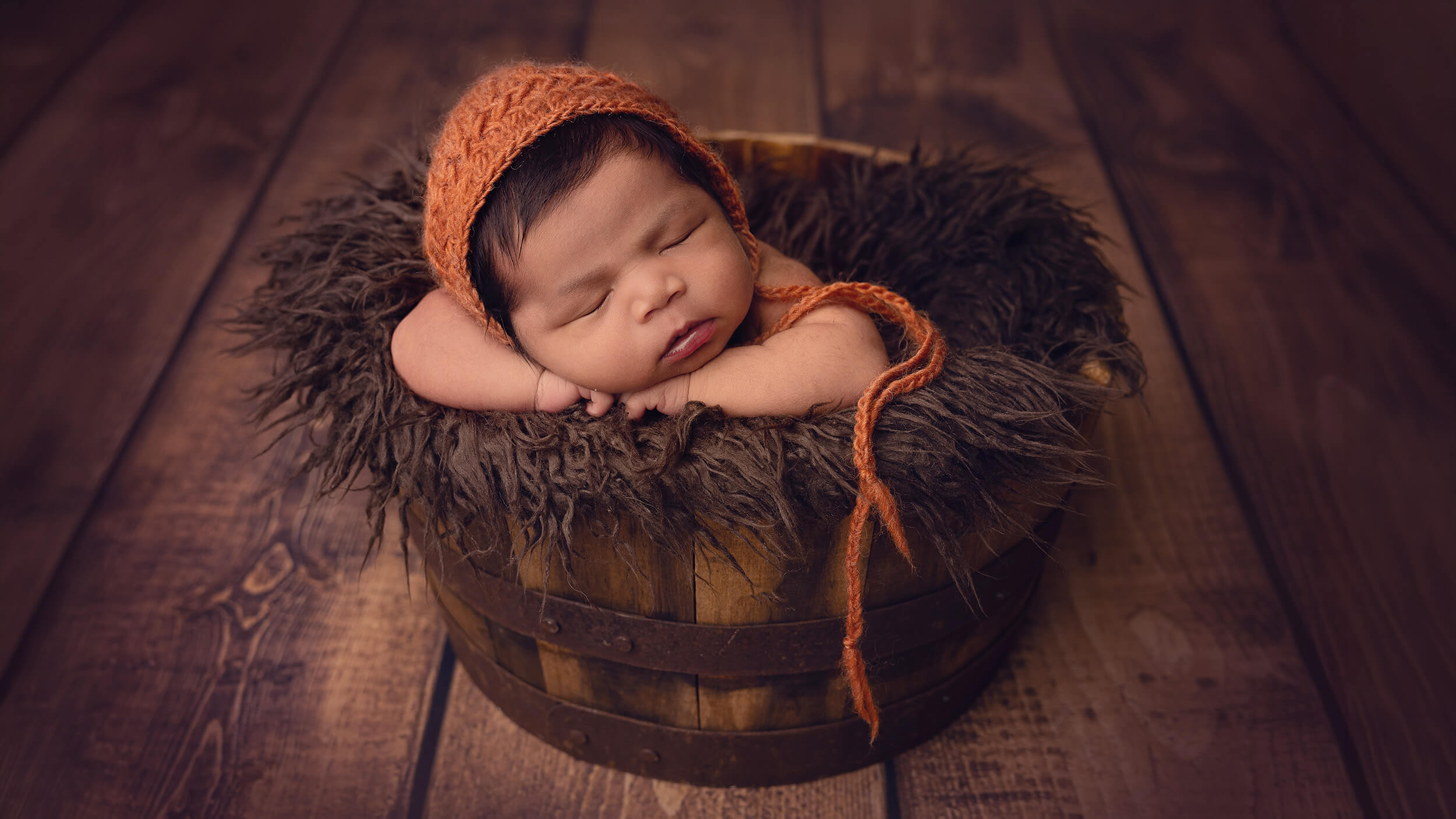 A newborn baby posed in a barrel, wearing a knitted orange hat at AnA Photography's studio in Cornwall.