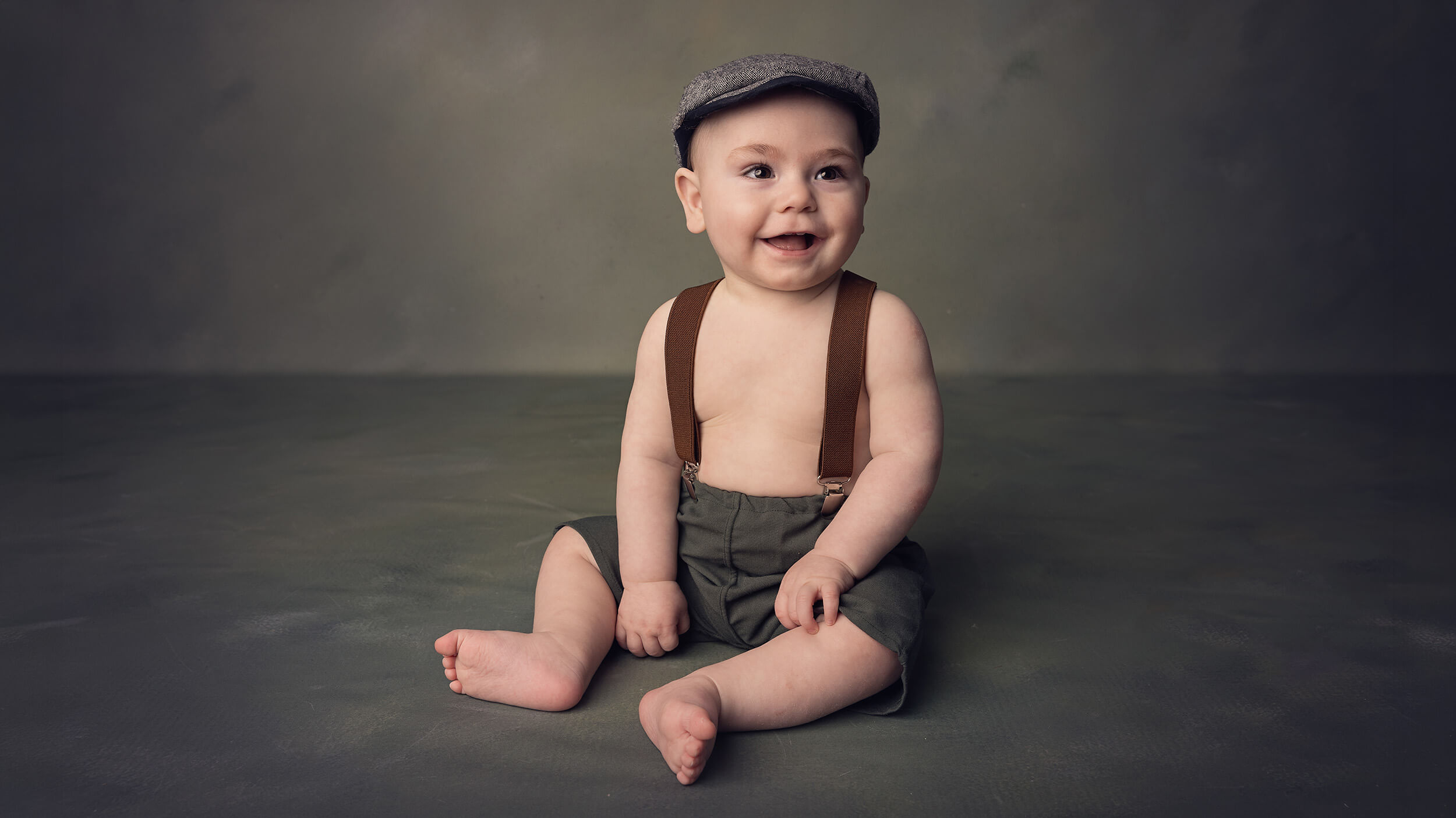 A toddler in a flat cap and dungarees posing for the camera at AnA Photography's studio in Cornwall.